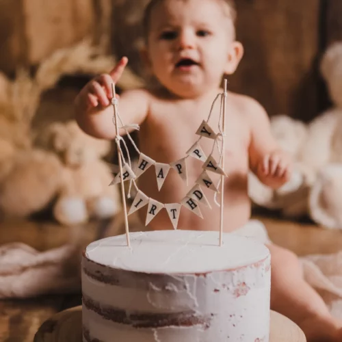 Bébé garçon curieux pointant du doigt son gâteau blanc avec guirlande Happy Birthday et ours en peluche - Studio professionnel proche Colombelles - Séance Anniversaire à Giberville.