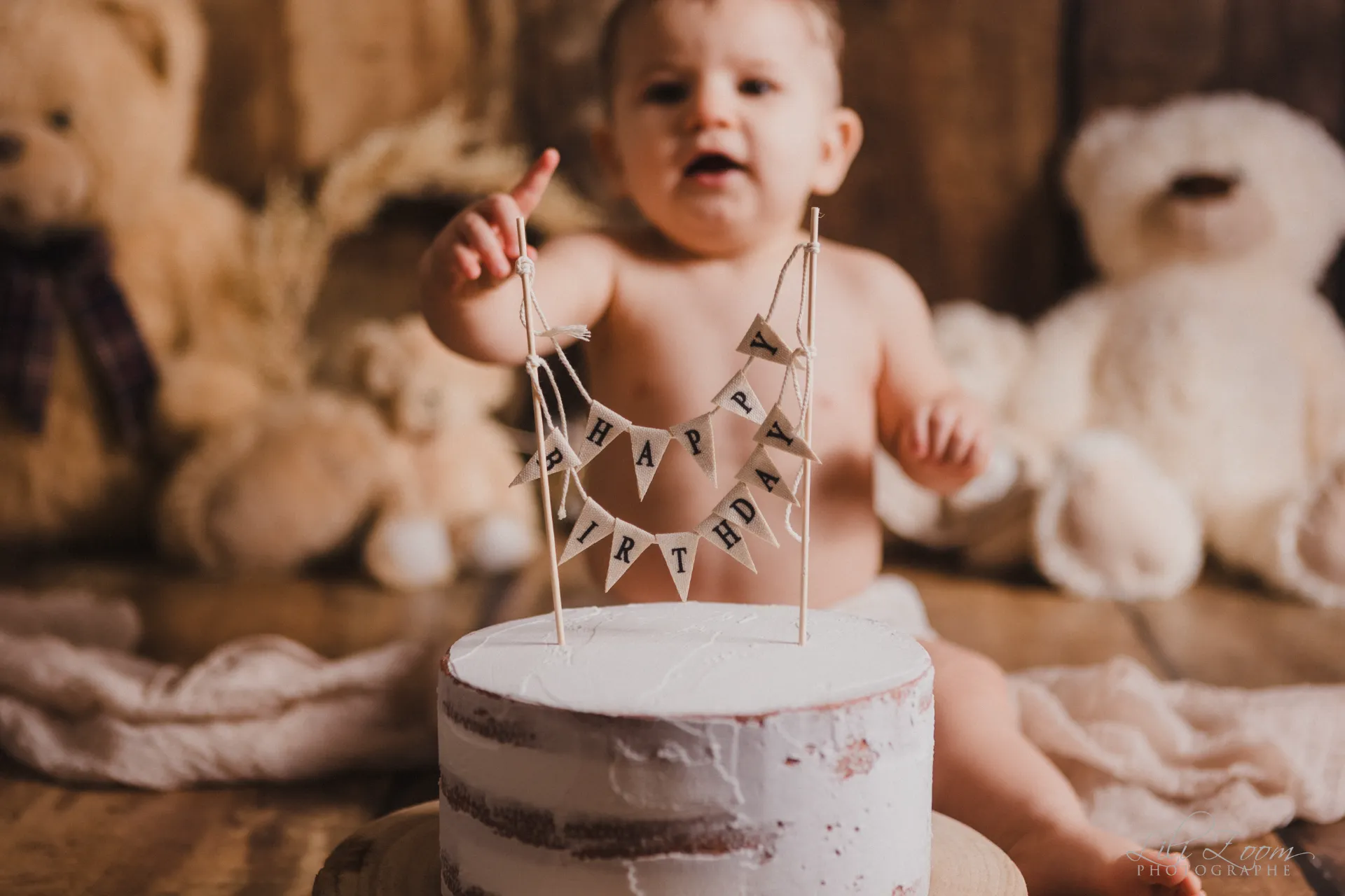 Bébé garçon curieux pointant du doigt son gâteau blanc avec guirlande Happy Birthday et ours en peluche - Studio professionnel proche Colombelles - Séance Anniversaire à Giberville.