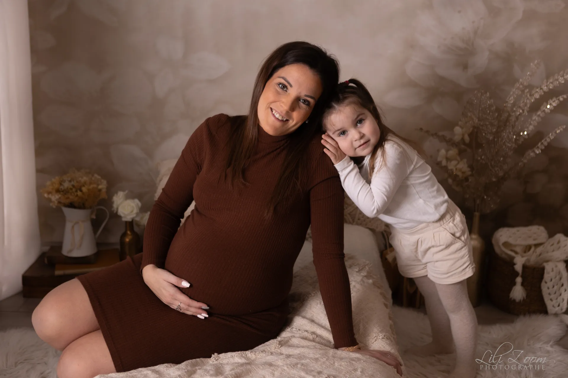 Une femme enceinte et sa petite fille sourient sur un lit dans une chambre bohème, séance grossesse en famille au studio de Giberville