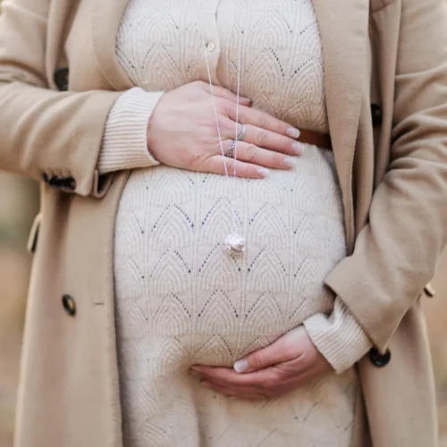 Femme enceinte en manteau de laine tient son ventre dans une forêt d'hiver, séance grossesse à Ifs