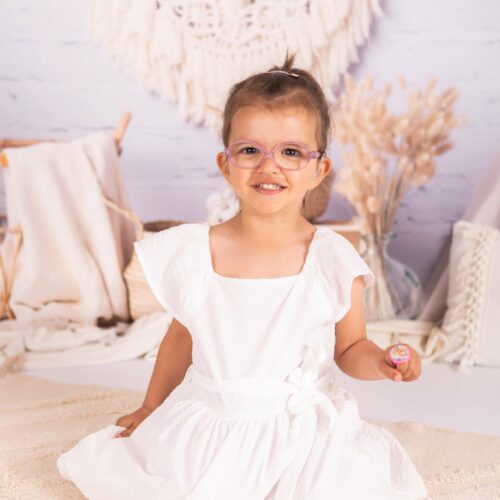 Une petite à lunettes portent une robe blanche sur un lit de dentelle de style bohème, séance enfant au studio à Caen