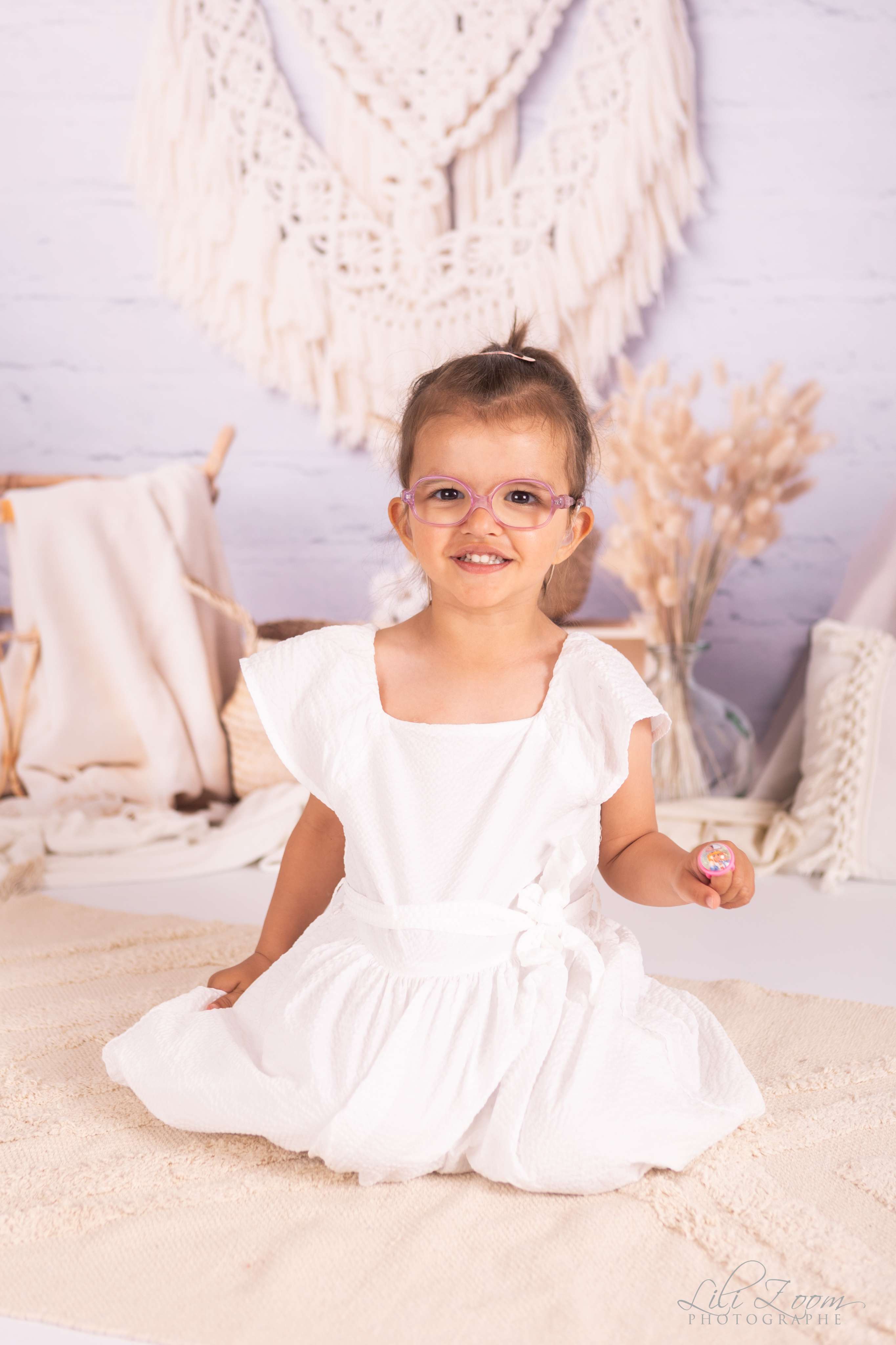 Une petite à lunettes portent une robe blanche sur un lit de dentelle de style bohème, séance enfant au studio à Caen