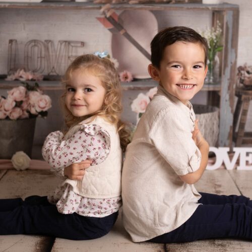 Une soeur et un frère sont dos à dos et sourient pendant une séance photographique en famille à Giberville