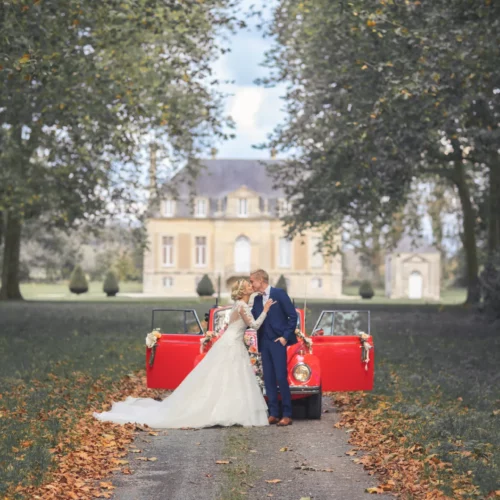 Couple de mariés s'embrassant tendrement devant une Coccinelle rouge vintage dans l'allée d'un château en automne - Photographe Famille à la Forêt de Bavent