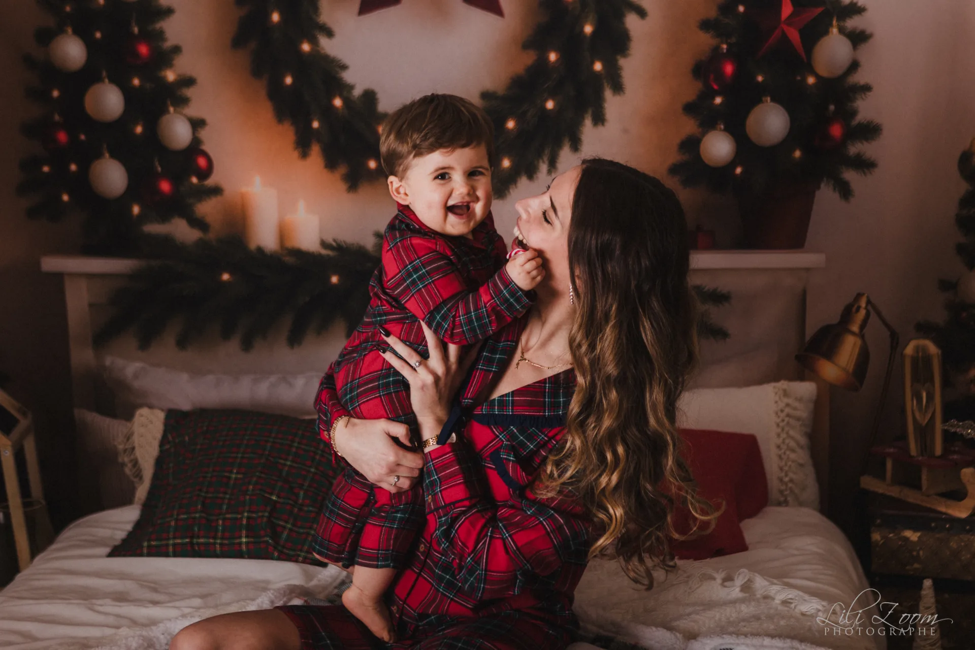 Une femme assise sur un lit tient un jeune enfant en pyjama à carreaux rouges et verts. Tous deux portent des tenues assorties, avec une décoration de Noël en arrière-plan.
