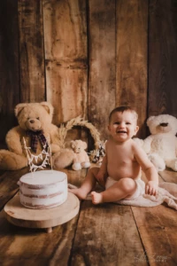 Bébé souriant devant son gâteau d'anniversaire entouré de peluches dans un décor en bois chaleureux - Photographe Bébé à Giberville Bébé souriant devant son gâteau d'anniversaire entouré de peluches dans un décor en bois chaleureux - Photographe Bébé à Giberville