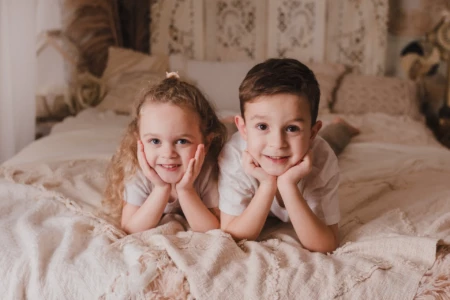 Frère et sœur complices souriant sur un lit dans un décor bohème et chaleureux - Photographe Famille à Giberville Frère et sœur complices souriant sur un lit dans un décor bohème et chaleureux - Photographe Famille à Giberville
