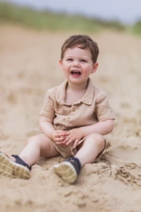 Petit garçon en tenue beige assis sur le sable riant aux éclats - Photographe enfant plage Merville-Franceville Petit garçon en tenue beige assis sur le sable riant aux éclats - Photographe enfant plage Merville-Franceville