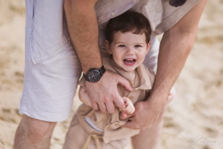 Petit garçon en tenue beige riant aux éclats dans les bras de son papa sur le sable - Photographe enfant Côte Fleurie Petit garçon en tenue beige riant aux éclats dans les bras de son papa sur le sable - Photographe enfant Côte Fleurie