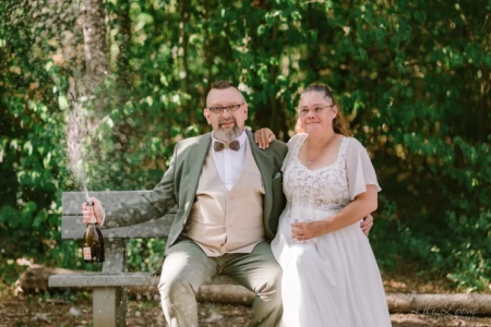 Couple de mariés assis sur un banc faisant sauter le bouchon de champagne dans un parc - Photographe mariage Courseulles-sur-Mer Couple de mariés assis sur un banc faisant sauter le bouchon de champagne dans un parc - Photographe mariage Courseulles-sur-Mer