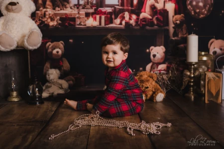 Un jeune enfant assis au milieu de peluches et de décorations. Un jeune enfant assis sur un sol en bois, portant un pyjama rouge à carreaux. Autour de lui se trouvent plusieurs peluches, des bougies et des objets décoratifs.
