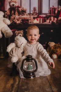 Jeune enfant assis au milieu d’ours en peluche et de décorations festives. Un jeune enfant assis sur un sol en bois tient un ours en peluche blanc, avec une petite lanterne décorative devant lui. Autour, plusieurs ours en peluche et des paquets cadeaux composent un décor festif.