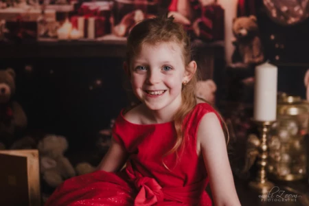 Portrait d’une enfant en robe rouge devant un décor sombre. Portrait d’une enfant en robe rouge, assise et souriante, devant un décor sombre avec une bougie et des objets décoratifs flous en arrière-plan.