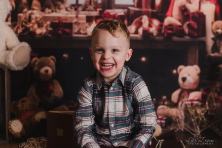 Un jeune enfant sourit devant un décor avec des ours en peluche. Un jeune enfant assis, souriant à l’objectif, devant un décor avec plusieurs ours en peluche et des lumières décoratives.