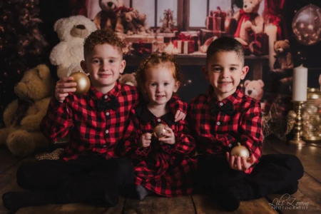 Trois enfants assis avec des boules décoratives dorées devant un décor de Noël. Trois enfants assis côte à côte sur un sol en bois, portant des vêtements à carreaux rouges et noirs, chacun tenant une boule décorative dorée. En arrière-plan, des peluches, des cadeaux et des décorations de Noël.