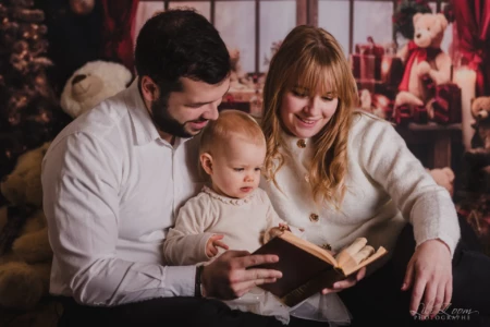 Un adulte et un enfant regardent un livre ouvert devant un décor avec des peluches et des cadeaux. Un homme, une femme et un jeune enfant sont assis ensemble et regardent un livre ouvert. L’arrière-plan est décoré de peluches et de cadeaux.