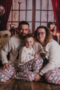 Trois personnes en pyjamas assortis devant un décor avec des ours en peluche. Une femme, un homme et un enfant sont assis sur le sol, en pyjamas assortis blancs à motifs hivernaux, devant un décor avec des ours en peluche et des rideaux rouges.