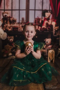 Petite fille en robe verte tenant une boule dorée devant un décor d’ours en peluche et de cadeaux. Une petite fille en robe verte est assise par terre et tient une boule dorée entre ses mains, devant un décor avec des ours en peluche et des cadeaux.